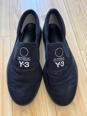 Svarta loafers Y-3 med tryck - Säljer ett par svarta loafers från Y-3 med vitt tryck på ovansidan där det står 'DIE MARKE MIT DEN 3 STREIFEN Y-3'. Skorna har en rund tå, elastiska paneler på sidorna och är tillverkade i textil och syntetmaterial.