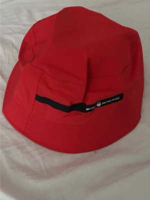 Röd bucket hat från Sail Racing - Snygg röd bucket hat från Sail Racing med svart detalj och logga framtill. Hatten är tillverkad i ett lätt och tåligt material, perfekt för soliga dagar. Insidan är fodrad i grått tyg för extra komfort.