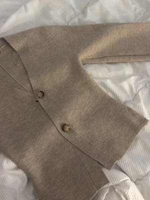 Beige kofta med stora knappar - Mysig beige kofta i mjukt stickat material med två stora, mönstrade knappar framtill och en knappdetalj vid ärmslutet. Koftan har v-ringning och lång ärm, perfekt för lager på lager och enkel att styla till olika outfits.