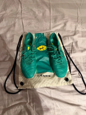 Nike Tiempo turkosa fotbollsskor - Säljer ett par Nike Tiempo fotbollsskor i en snygg turkos färg med gula detaljer. Skorna har klassisk snörning, dobbar för spel på gräs och är tillverkade i syntetmaterial. Perfekta för dig som vill ha bra touch och kontroll på planen. Medföljer originalpåse.