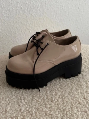 Beige chunky loafers med platåsula - Hade på mig en gång. Säljer ett par beige loafers med glansig finish och kraftig svart platåsula. Skorna har snörning med svarta skosnören och grov sula för extra attityd. Perfekta för dig som vill sticka ut och addera höjd till din stil.