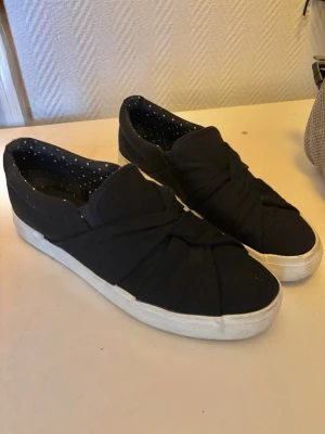 Svarta skor - Stilrena svarta slip-on sneakers med en snygg knutdetalj på ovansidan. Tillverkade i tygmaterial och passar perfekt för både avslappnad och uppklädd look.