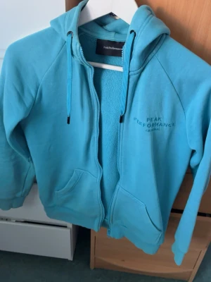  Peak Performance hoodie - Snygg turkos hoodie från Peak Performance med dragkedja, två fickor framtill och broderad logga på bröstet. Mjuk insida och justerbar huva med snören. Perfekt för chill dagar eller när du vill ha en sportig look.