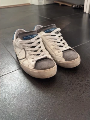Philippe Model vita och grå sneakers - Snygga sneakers från Philippe Model i vitt och grått med blå detaljer. Skorna har lågt skaft, vita skosnören och en klassisk siluett. Ovandelen är i läder och mocka vilket ger en schysst kontrast. Perfekta för dig som gillar stilrena och trendiga sneakers.