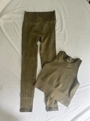 Olivgrönt ribbat träningsset  - Säljer ett olivgrönt ribbat träningsset från STRONGER. Setet består av ett par höga tights och en matchande crop top med bred resår nedtill. Båda delarna är i stretchigt material och har en sportig look. Perfekt för gymmet eller chill dagar.
