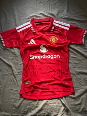 Manchester United 26/27 - Säljer en röd Manchester United matchtröja från Adidas med klubbmärke på bröstet och Snapdragon-logga framtill. Tröjan har vita detaljer på axlar och krage, samt korta ärmar. Tillverkad i lätt och ventilerande funktionsmaterial, perfekt för fotboll.