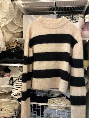 Beige och svart randig stickad tröja - Stickad tröja med breda svarta och beige ränder. Tröjan har en rak, något boxig passform, ribbad rund halsringning samt ribbade muddar vid ärmslut och nederkant. Materialet ser mjukt och fluffigt ut, perfekt för kyligare dagar.