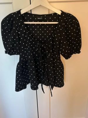 Svart prickig blus med knyt - Supersöt svart blus med vita prickar och puffärmar. Djup v-ringning med knyt framtill och volang nedtill. 