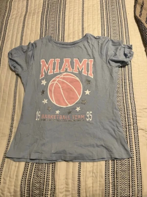 Blå t-shirt med MIAMI-tryck - Ljusblå t-shirt från Primark med rosa och vitt MIAMI-tryck och basketbollmotiv framtill. T-shirten har korta ärmar och glittriga silverdetaljer runt texten och stjärnorna. Perfekt för en sportig och avslappnad stil.