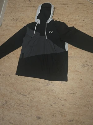 Svart och grå vindjacka Under Armour - Snygg vindjacka från Under Armour i svart och grått med vit logga på bröstet och stor grafisk print på ryggen. Jackan har huva med dragsko och långa ärmar. Tillverkad i lätt och vindtätt material, perfekt för blåsiga dagar.