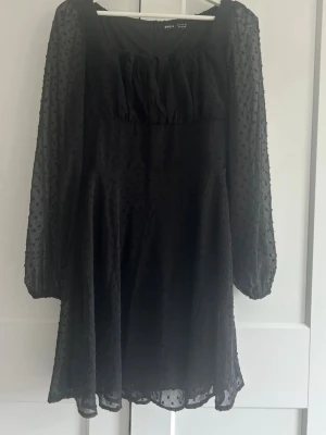 Svart kortklänning med prickigt mönster - Snygg svart kortklänning med transparenta långärmar och diskret prickigt mönster. Klänningen har en rynkad detalj vid bysten och är tillverkad i ett lätt, luftigt material som ger en elegant look. Perfekt för dig som vill ha något stilrent och trendigt.