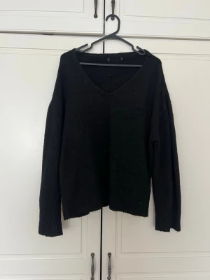 Svart stickad tröja med v-ringning - En enkel och stilren svart stickad tröja med djup v-ringning och långa ärmar. Tröjan har ribbade muddar vid ärmslut och nederkant, vilket ger en snygg och avslappnad look. Perfekt att matcha med jeans eller kjol för en trendig outfit.