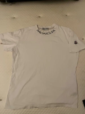 Vit Moncler t-shirt med logga - Vit t-shirt från Moncler med svart logotyptext runt halsen och klassisk Moncler-patch på ärmen. T-shirten har en enkel och stilren design med korta ärmar och rund hals. Perfekt för dig som gillar exklusiva streetwear-plagg.