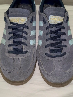 Adidas Spezial blå sneakers - Snygga Adidas Spezial sneakers i blå mocka med ljusblå ränder och mörkblå snören. Skorna har klassisk Adidas-design med gummisula och guldfärgade detaljer på sidan och tungan. Perfekta för dig som gillar retrostil och streetwear.