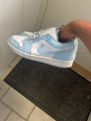 Nike Air Jordan 1 Low UNC blå/vit - Säljer ett par Nike Air Jordan 1 Low i färgen UNC blå och vit. Skorna har klassisk låg siluett, perforerad tåbox och ikoniska Swoosh-loggan på sidan. Tillverkade i läder med matchande blå skosnören och Jumpman-logga på plösen.
