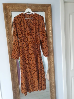 Leopardmönstrad omlottklänning - Snygg långklänning i leopardmönster med omlottmodell och knyt i midjan. Klänningen har långa ärmar och en varm brun ton som sticker ut. Perfekt för dig som vill ha en trendig och bekväm look. Mer som 40 i syorlek