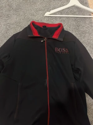 Svart zip hoodie från Hugo Boss - Snygg svart hoodie från Hugo Boss med röd dragkedja och röda detaljer på kragen där det står 'HUGO BOSS'. Stort BOSS-tryck på bröstet. Tillverkad i mjukt material och har två fickor framtill. Perfekt för en stilren och sportig look.