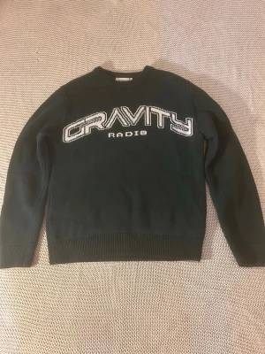 Mörkgrön stickad tröja GRAVITY - Mörkgrön stickad tröja med texten 'GRAVITY RADIO' i vitt och svart på bröstet. Tröjan har rund halsringning, ribbade muddar och är gjord i ett tjockare stickat material. Perfekt för kalla dagar och en avslappnad streetwear-look.