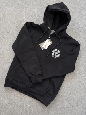 Chrome Hearts Hoodie - Snygg svart hoodie från Chrome Hearts med stort vitt tryck på ryggen och mindre logga på bröstet. Klassisk känguruficka framtill, dragsko i huvan och ribbade muddar. Perfekt för dig som gillar streetwear och vill sticka ut.