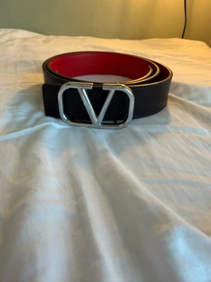 Valentino bälte - Snyggt svart bälte med ett stort, silverfärgat V-format spänne och röd insida. Perfekt accessoar för att lyfta vilken outfit som helst. Bältet har en stilren och modern design med bred rem.