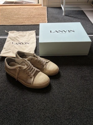 Beige låga sneakers från Lanvin - Snygga beige sneakers från Lanvin med klassisk låg siluett. Skorna har rund tå, matchande snörning och detaljer i både läder och textil. Kommer med originalkartong och dustbag. Perfekta för dig som gillar stilrena och tidlösa sneakers.