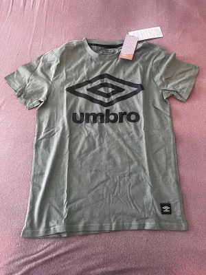 Grön t-shirt från Umbro med logga - Snygg grön t-shirt från Umbro med stor svart logga på bröstet. Klassisk passform och rund halsringning. Tillverkad i mjuk bomull som känns skön mot huden. Perfekt för dig som gillar sportiga och stilrena plagg.