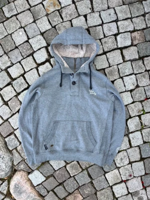 Grå hoodie med knappar och teddyfoder - Mysig grå hoodie med teddyfoder i huvan och två knappar vid halsen. Har en stor magficka, svarta dragsnören och broderad text på bröstet. Ribbstickade muddar vid ärmar och nederkant. Perfekt för chill dagar och streetwear.