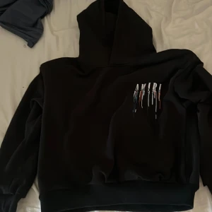 Svart Amiri hoodie med färgstänk - Svart hoodie från Amiri med stor huva och coolt tryck med färgstänk och logga både fram och bak. Oversized fit med ribbade muddar. Perfekt för dig som vill sticka ut med streetstyle och exklusiv känsla. (PASSAR FOLK SOM ÄR 155-157!)