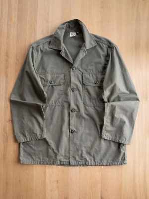Orslow Fatigue Shirt, size 3/L - Stilren olivgrön overshirt i bomull med klassisk krage och fyra knappar framtill. Två bröstfickor med lock och knapp ger en cool utility-look. Perfekt att bära öppen eller stängd över t-shirt. Rak passform och avslappnad stil.