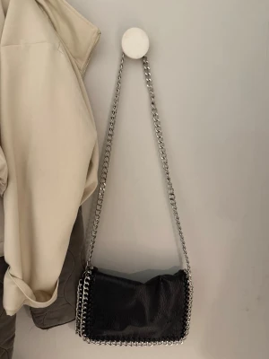 Svart montini axelväska med kedjerem - Snygg svart axelväska i fejkläder med silverfärgad kedjerem och detaljer. Väskan har en dragkedja på baksidan och dekorativa kedjor längs kanterna. Perfekt för dig som gillar en edgy och trendig stil.