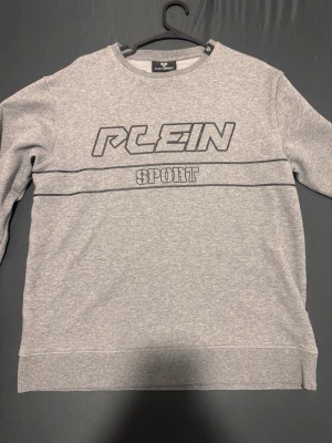 Grå sweatshirt från Plein Sport - Snygg grå sweatshirt från Plein Sport med tryckt logga och text på bröstet. Tröjan har rund halsringning och raka ärmar. Perfekt för en sportig och avslappnad stil. Materialet känns mjukt och bekvämt mot huden.