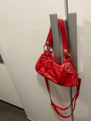Röd handväska från Marina Galanti - Cool röd handväska från Marina Galanti med stora silverfärgade ringdetaljer och två axelband. Väskan har dragkedja upptill och en beige innerficka med logga. Materialet ser ut som glansigt konstläder. Perfekt för dig som vill sticka ut med färg!