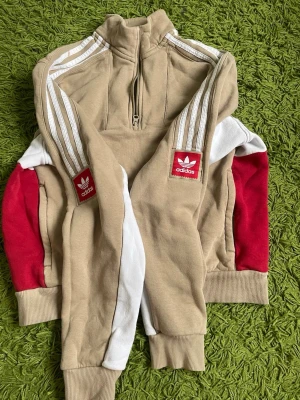 Beige Adidas hoodie med röd detalj - Säljer en beige hoodie från Adidas med halv dragkedja, vita ränder på axlarna och röda samt vita detaljer på ärmarna. Klassisk Adidas-logga på ärmen. Mjuk bomullskänsla och ribbade muddar. Perfekt för en sportig och avslappnad stil.