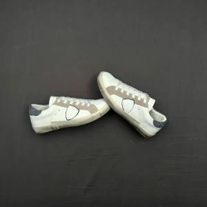 Vita Philippe Model skor med ljusblåa detalj  - Snygga Philippe Model skor i vitt och beige med ljusblå mockadetalj på hälen. Skorna har klassisk låg profil, vita snören och en beige sula. Materialet är en mix av slätt läder och mocka, vilket ger en stilren och trendig look.