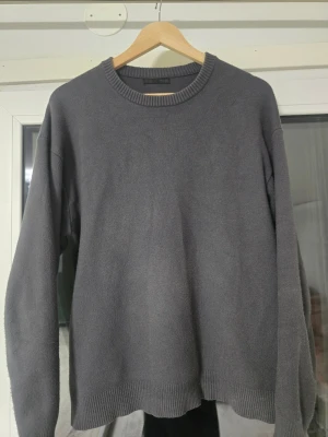 Mörkgrå stickad tröja från Zara - En enkel och stilren mörkgrå stickad tröja från Zara. Tröjan har rund halsringning, långa ärmar och ribbade muddar vid hals, ärmslut och nederkant. Perfekt för lager på lager och passar till det mesta i garderoben.