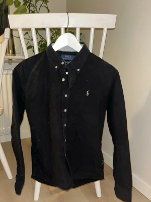 Svart skjorta från Polo Ralph Lauren - Svart långärmad skjorta från Polo Ralph Lauren med klassisk button-down krage och broderad logga på bröstet. Skjortan har vita knappar och är i en normal passform. Perfekt för en stilren look.
