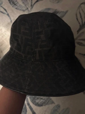 Svart bucket hat från Fendi - Snygg svart bucket hat från Fendi med det klassiska FF-mönstret över hela hatten. Tillverkad i ett mjukt tyg med diskret glans och fodrad insida för extra komfort. Perfekt accessoar för att lyfta din streetwear-look.
