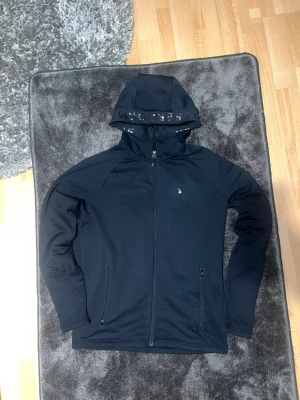 Svart hoodie peak performance hoddie - Snygg svart hoodie med hel dragkedja framtill och två fickor med dragkedja. Huva med textdetalj och minimalistisk logga på bröstet. Tillverkad i ett mjukt och stretchigt material som passar perfekt till en avslappnad stil.M men kan passa S 