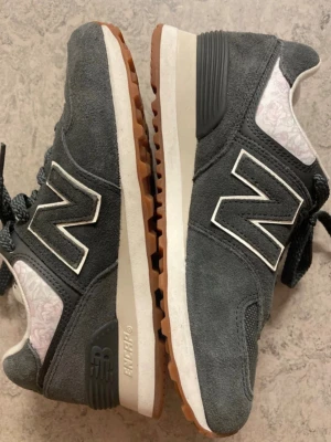 New Balance 574 grå sneakers - Grå New Balance 574 sneakers i mocka med vita och svarta detaljer. Klassisk N-logga på sidan, vit mellansula och gummifärgad yttersula. Insidan har ett diskret blommönster i ljusrosa och vitt. Skön och tidlös modell med snörning.storlek 36,5