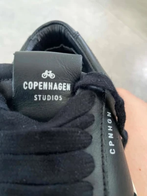 Svarta sneakers från Copenhagen Studios - Snygga svarta sneakers från Copenhagen Studios med vita sulor och svarta skosnören. Skorna har detaljer i mocka och läder, samt texten CPNHGN på sidan. Klassisk låg modell med perforerade partier för extra stil.