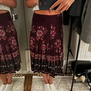 Mörkröd blommig långkjol - Säljer en mörkröd långkjol med blommigt mönster i rosa och vitt. Kjolen har en lätt utställd form och ett dekorativt band nertill. Perfekt för dig som vill ha en bohemisk och färgstark stil.