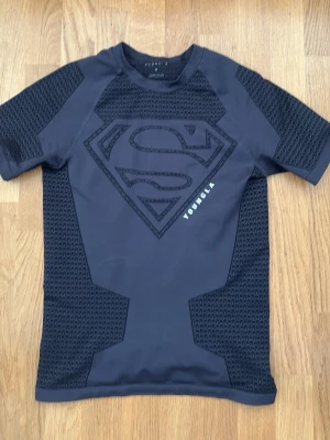 Superman YoungLA - Snygg svart kompressionströja från YoungLA med Superman-logga på bröstet och detaljerade mönster på ärmar och sidor. Tillverkad i stretchigt funktionsmaterial som passar perfekt till träning eller gym. Kortärmad modell med tight passform.
