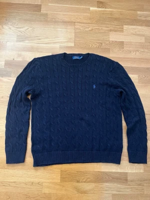 Marinblå kabelstickad Ralph Lauren tröja i storlek XXL - Kabelstickad marinblå tröja från Polo Ralph Lauren med rund halsringning och broderad logga på bröstet. Tröjan har långa ärmar och ribbade muddar vid ärmslut och nederkant. Perfekt för lager på lager och klassisk stil. Tröjan är som ny och det finns inga tecken på användning eller slitage. 
