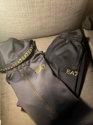 Svart EA7 set med guldtryck - Säljer en svart tracksuit från EA7 Emporio Armani med guldfärgade loggor på både hoodie och byxor. Hoodien har dragkedja, huva med snörning och tryck längs ärmen. Byxorna har resår i midjan och sidofickor. Perfekt för en sportig och trendig look.