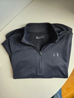 Svart half-zip tröja från Under Armour - Svart half-zip tröja från Under Armour med hög krage och diskret logga på bröstet. Tillverkad i mjukt och värmande material med ColdGear-teknologi. Perfekt för kyliga dagar och sportiga outfits.