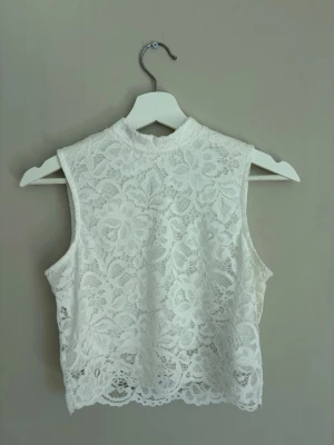 Vit spetsärmlös topp med hög krage - Säljer en vit ärmlös topp i spets med hög krage och vackert blommigt mönster. Toppen har en liten knapp i nacken och är croppad i modellen. Perfekt för dig som gillar romantisk och trendig stil.