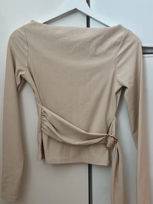 Beige omlottopp med bälte - Trendig beige omlottopp med lång ärm och snyggt bälte i midjan med guldfärgat spänne. Toppen har en stilren och enkel design som passar perfekt till många olika looks. Tillverkad i mjukt material för en bekväm känsla.