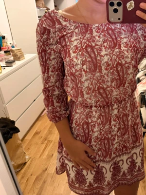 Rödvit mönstrad kortklänning - Kort klänning med rödvitt paisleyinspirerat mönster och trekvartsärm. Klänningen har en markerad midja och rund halsringning. Perfekt för sommarens varma dagar. Den har även öppen rygg, medella för att få fler bilder