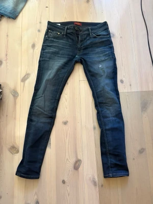 Jack & Jones  - Snygga mörkblå slim fit jeans med lätt slitningar och klassisk femficksdesign. Jeansen har en smal passform och subtila tvättade detaljer på låren och knäna. Perfekta för en trendig och avslappnad look. Storlek 30/30