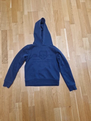 Mörkblå hoodie från GANT - Snygg mörkblå hoodie från GANT med broderad logga och texten 'American Sportswear' på bröstet. Klassisk huva och ribbade muddar vid ärmslut och nederkant. Perfekt för en avslappnad och sportig stil.